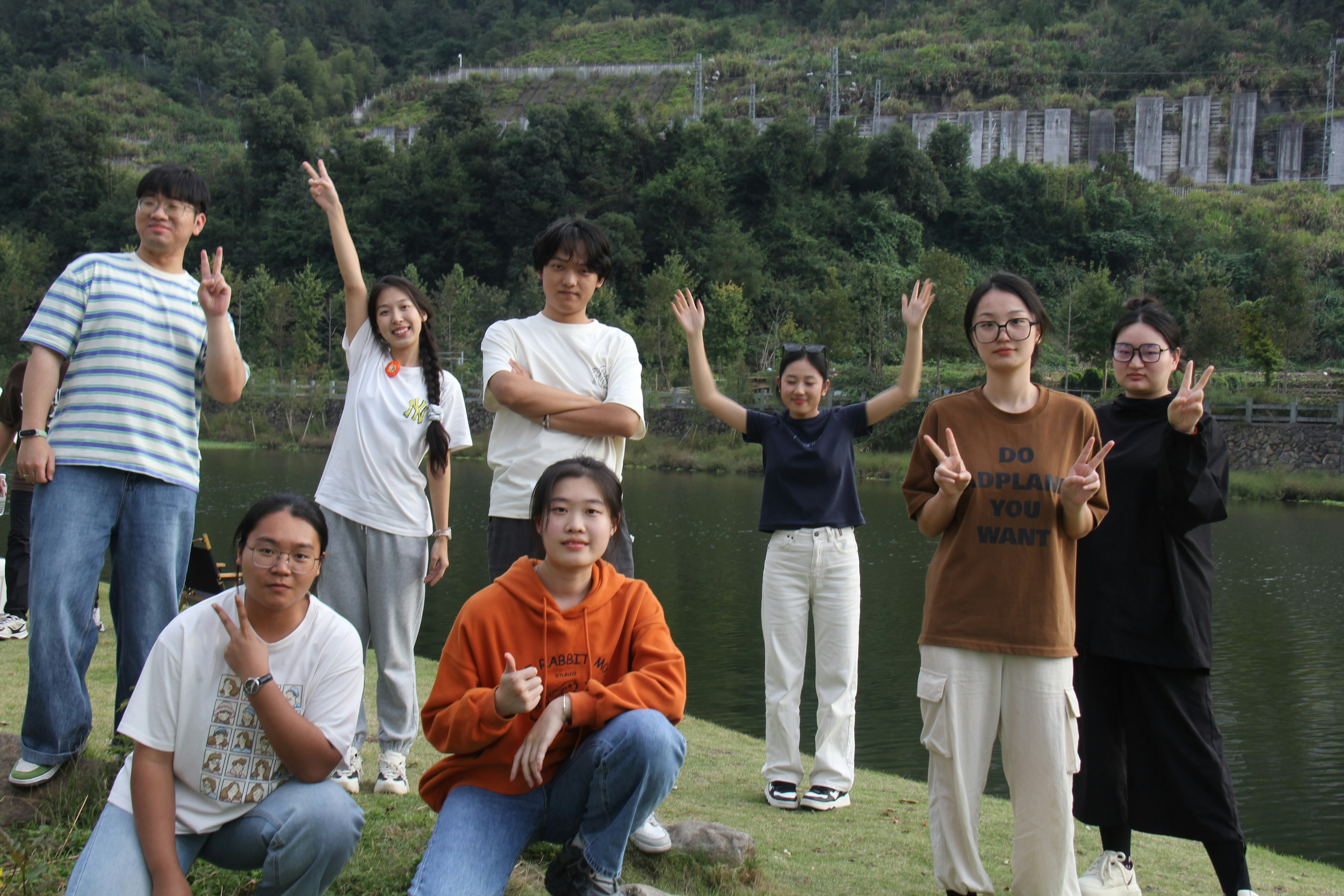 Eight young people pose outdoors near a lake and mountains